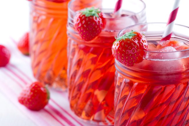 Glasses of red strawberry juice with frozen icecubes and striped straws on vintage towel and wooden background
